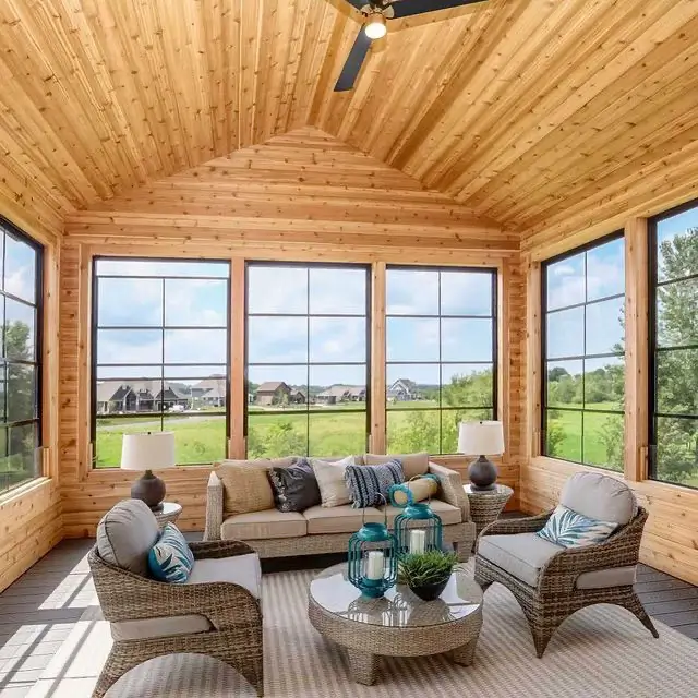 Interior of a sunroom with wood-paneled walls and ceiling, featuring Sunspace WeatherMaster windows and wicker lounge furniture.